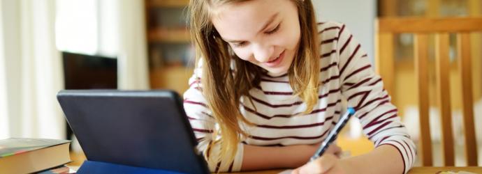 young girl sitting at a table colouring on a piece of paper with a tablet device in front of her