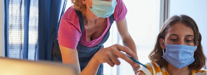An image of a teacher wearing a face covering whilst helping a student who is also wearing a face covering