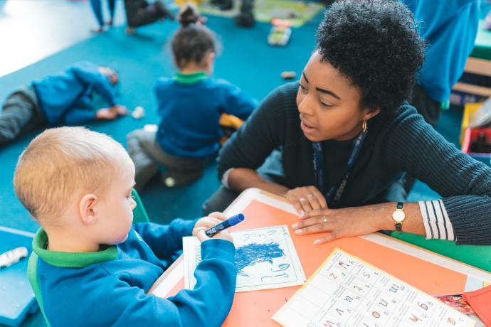 Image shows a teacher talking to a primary aged pupil who is colouring in.